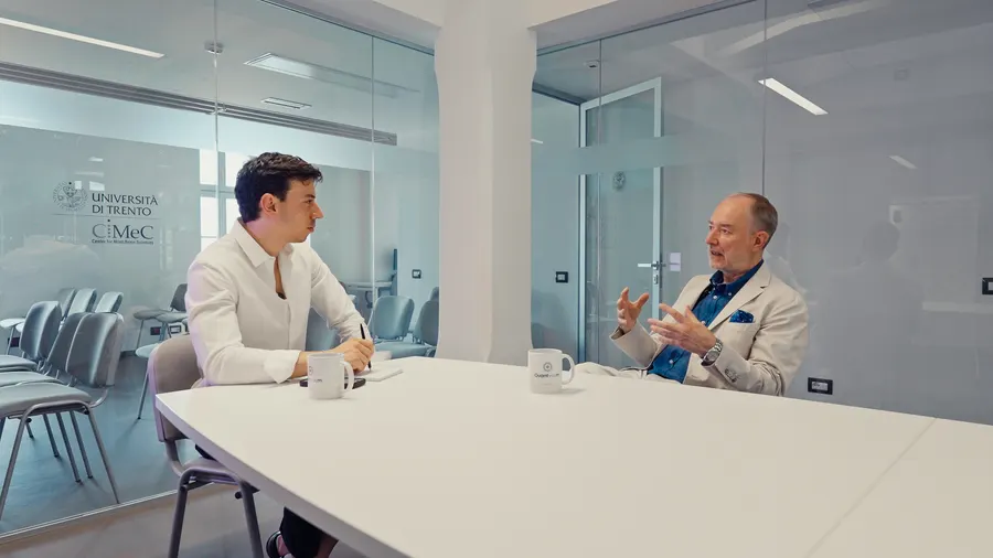 La sala riunioni vista di lato: l'intervistatore e l'ospite seduti ai due capi di un lungo tavolo bianco, con due tazze Quantyoum davanti.