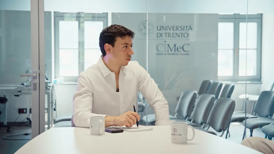 L'intervistatore ascolta prendendo appunti su un taccuino, davanti alla vetrata con il logo del CIMeC dell'Università di Trento.